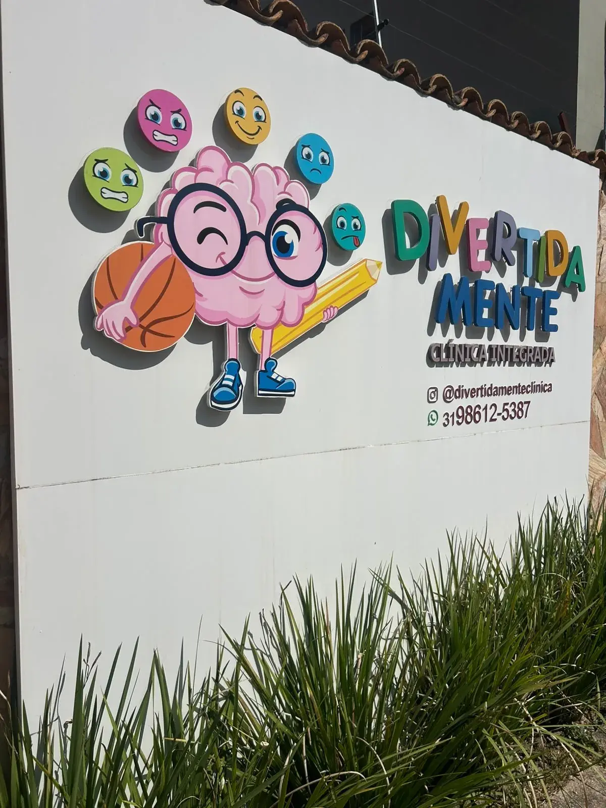 Fachada da clínica Divertida Mente com o letreiro colorido, o mascote de cérebro de óculos e carinhas de emoções na parede branca